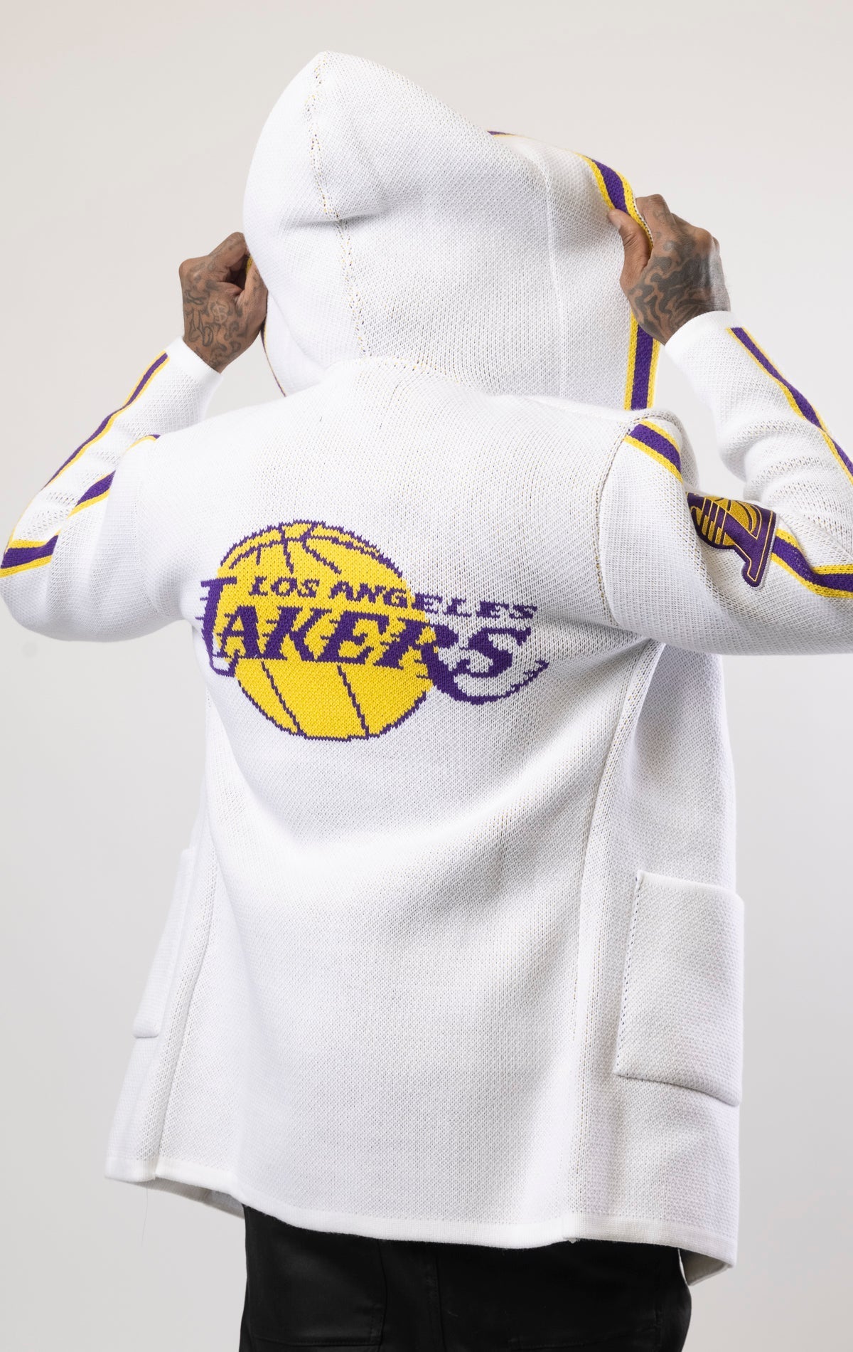 White LA LAKERS Cardigan Sweater. This cozy jacket is designed with a stylish three-quarter length and boasts bold team branding on the front, back, and sleeve. Complete with a full-zip closure, attached hood, and front pockets, this knit sweater offers b