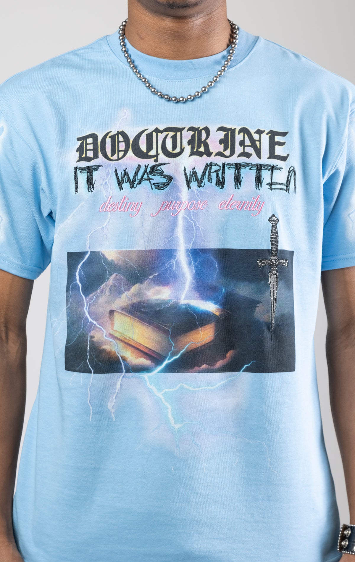 Sky blue t-shirt with a digital photo print on the front and back, embroidered text on the front, and a 3D metallic dagger embroidered on the back.