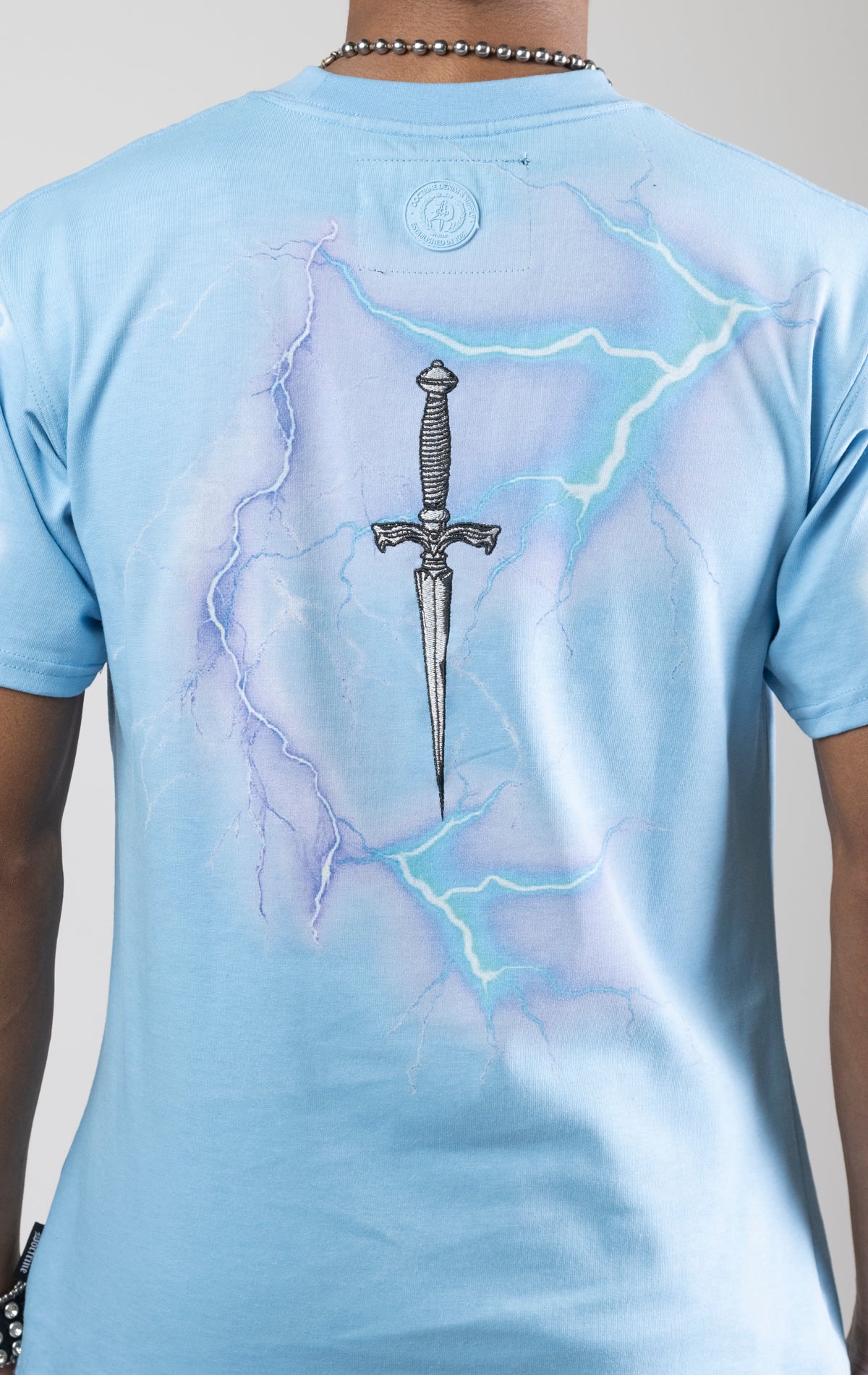 Sky blue t-shirt with a digital photo print on the front and back, embroidered text on the front, and a 3D metallic dagger embroidered on the back.