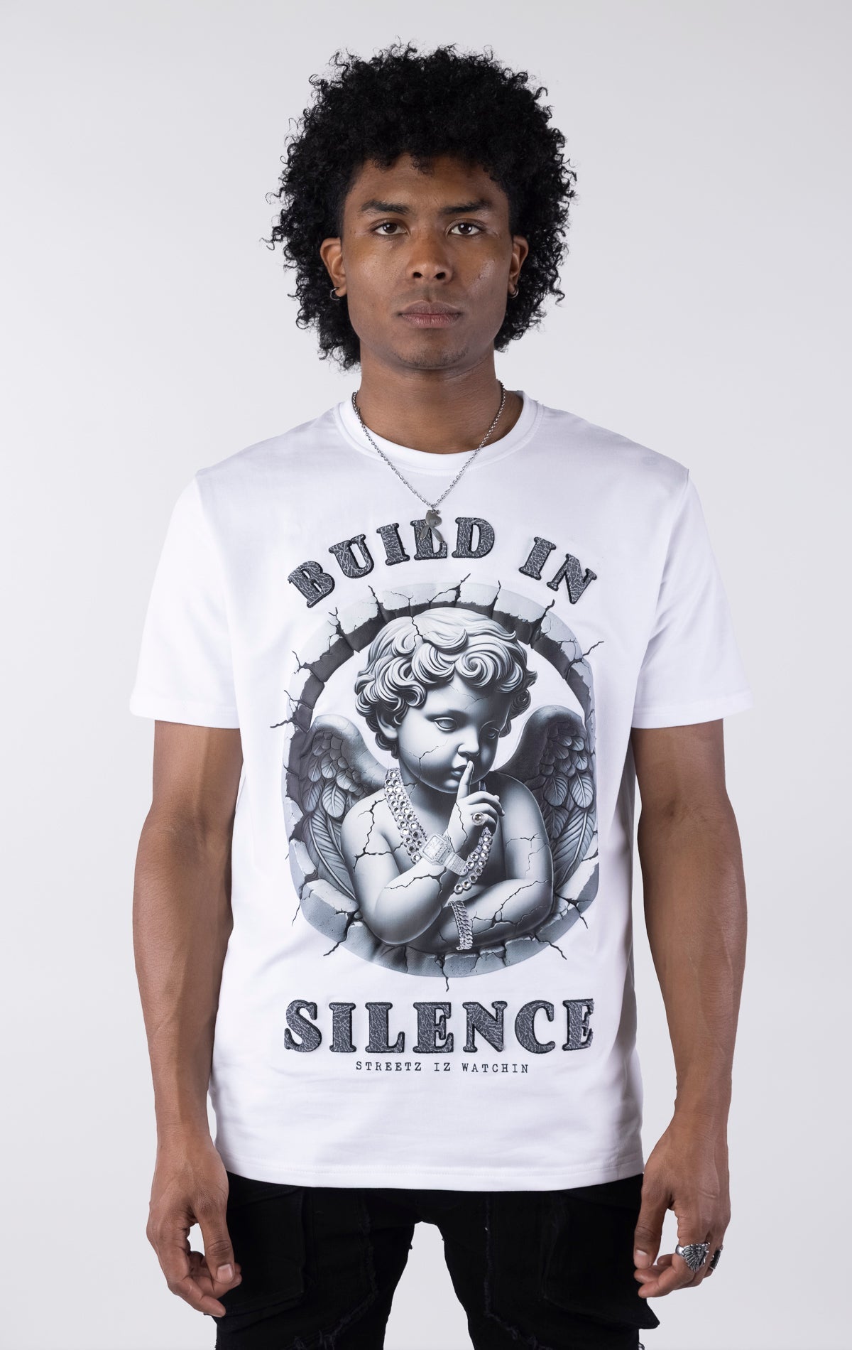 White t-shirt with a large graphic of a monochromatic cherub statue wearing chains and holding a finger to its lips. The graphic is surrounded by cracked stone and the text "BUILD IN SILENCE" is printed above and below in a textured black font.