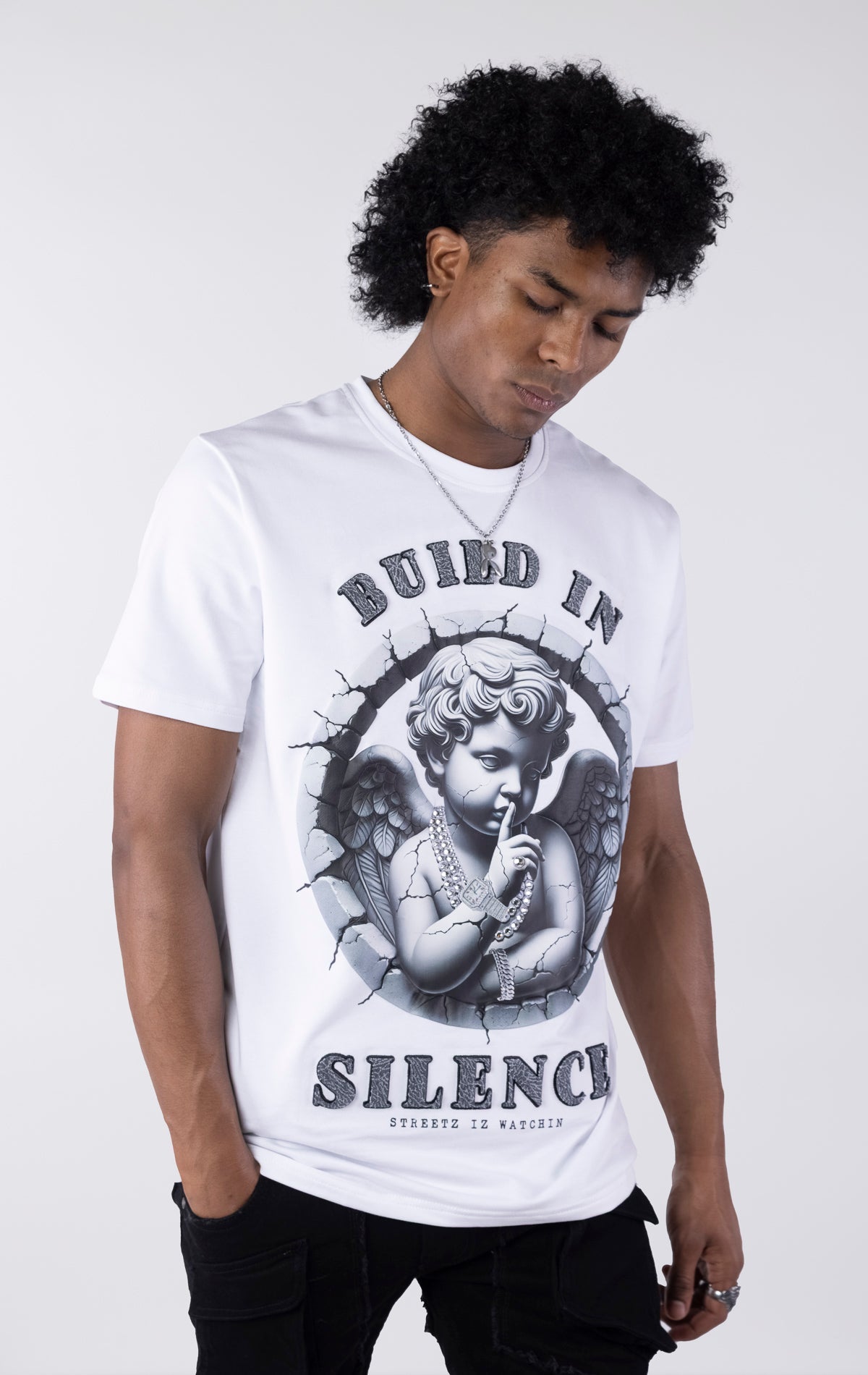 White t-shirt with a large graphic of a monochromatic cherub statue wearing chains and holding a finger to its lips. The graphic is surrounded by cracked stone and the text "BUILD IN SILENCE" is printed above and below in a textured black font.