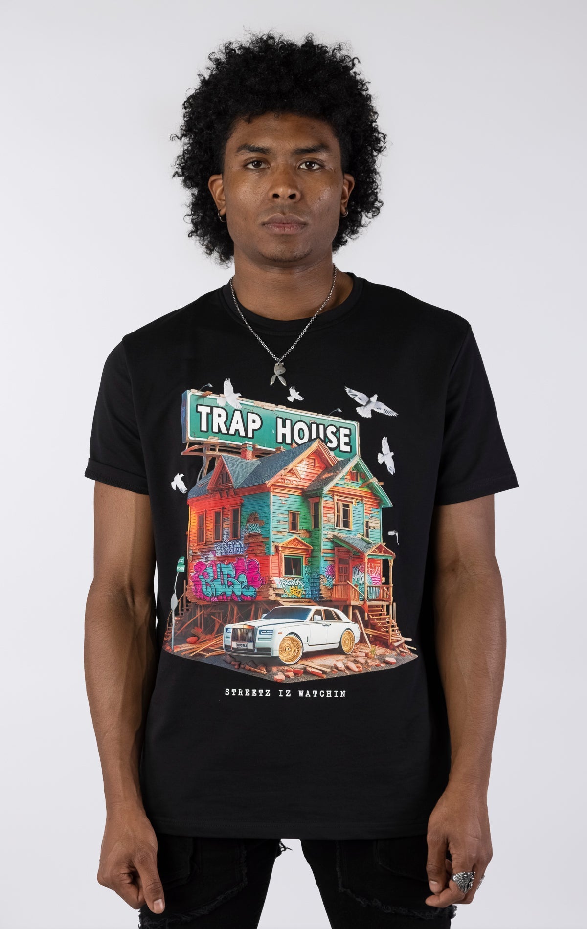 Black t-shirt with a large, colorful graphic of a run-down house with graffiti and a "TRAP HOUSE" sign on top. A white luxury car with gold rims is parked in front. White birds are flying around the house.