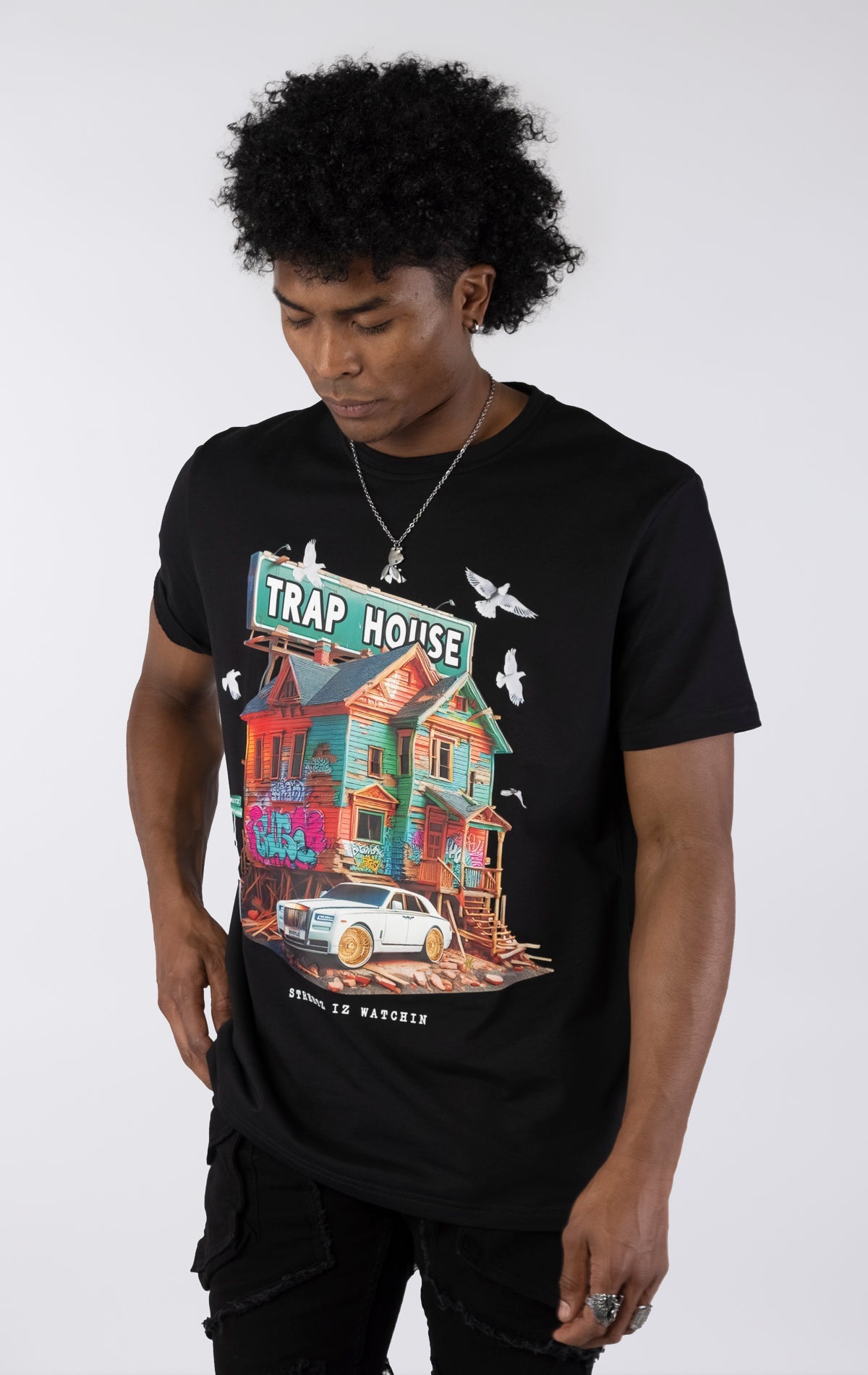 Black t-shirt with a large, colorful graphic of a run-down house with graffiti and a "TRAP HOUSE" sign on top. A white luxury car with gold rims is parked in front. White birds are flying around the house.