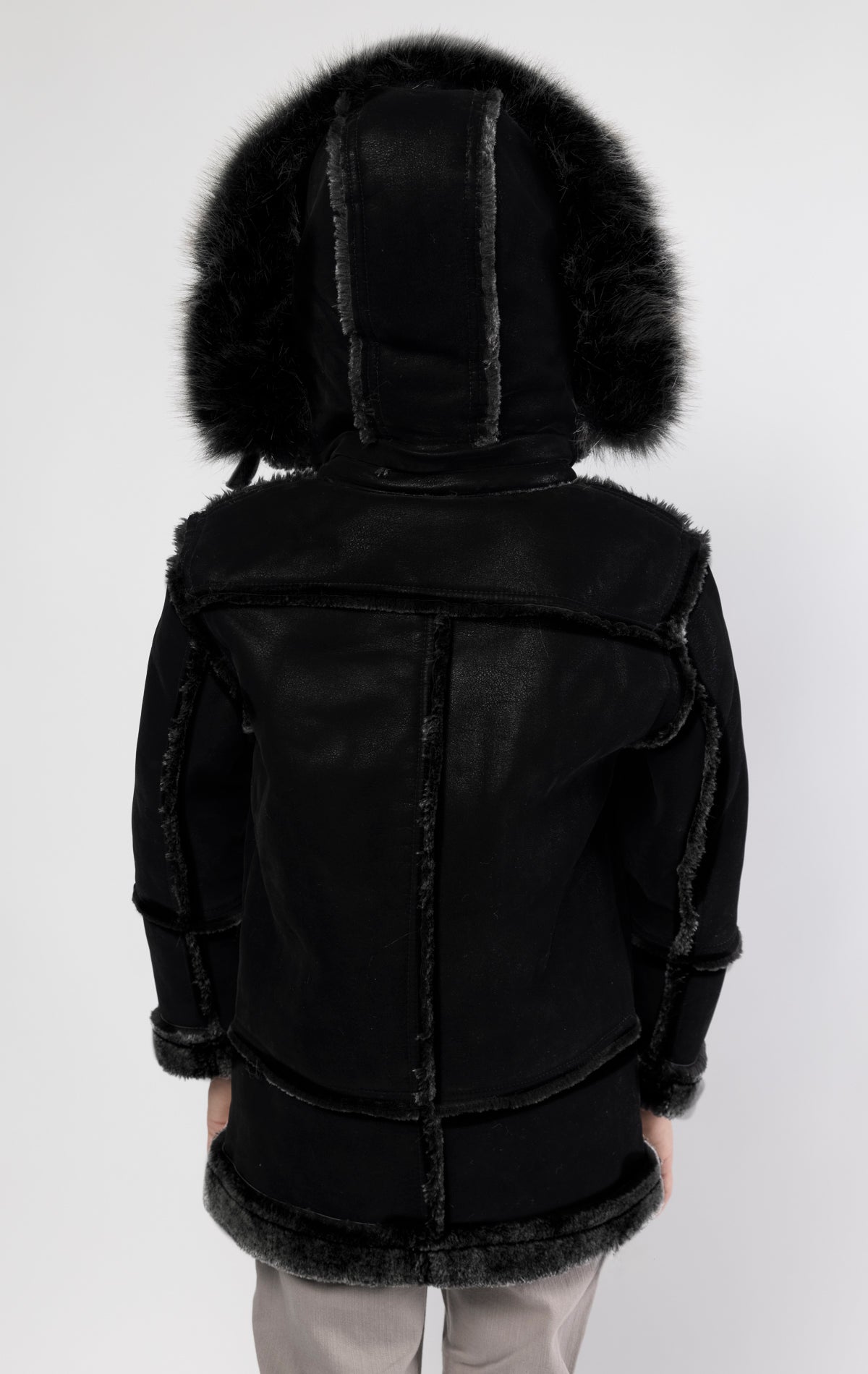 Black jacket lined with plush faux shearling throughout the body and sleeves. It also features shearling-accented pockets, a horn toggle front closure, and vegan suede straps with a buckle at the neck. For added convenience, a detachable faux fox fur hood with a zipper is included. This product is also available for adults.