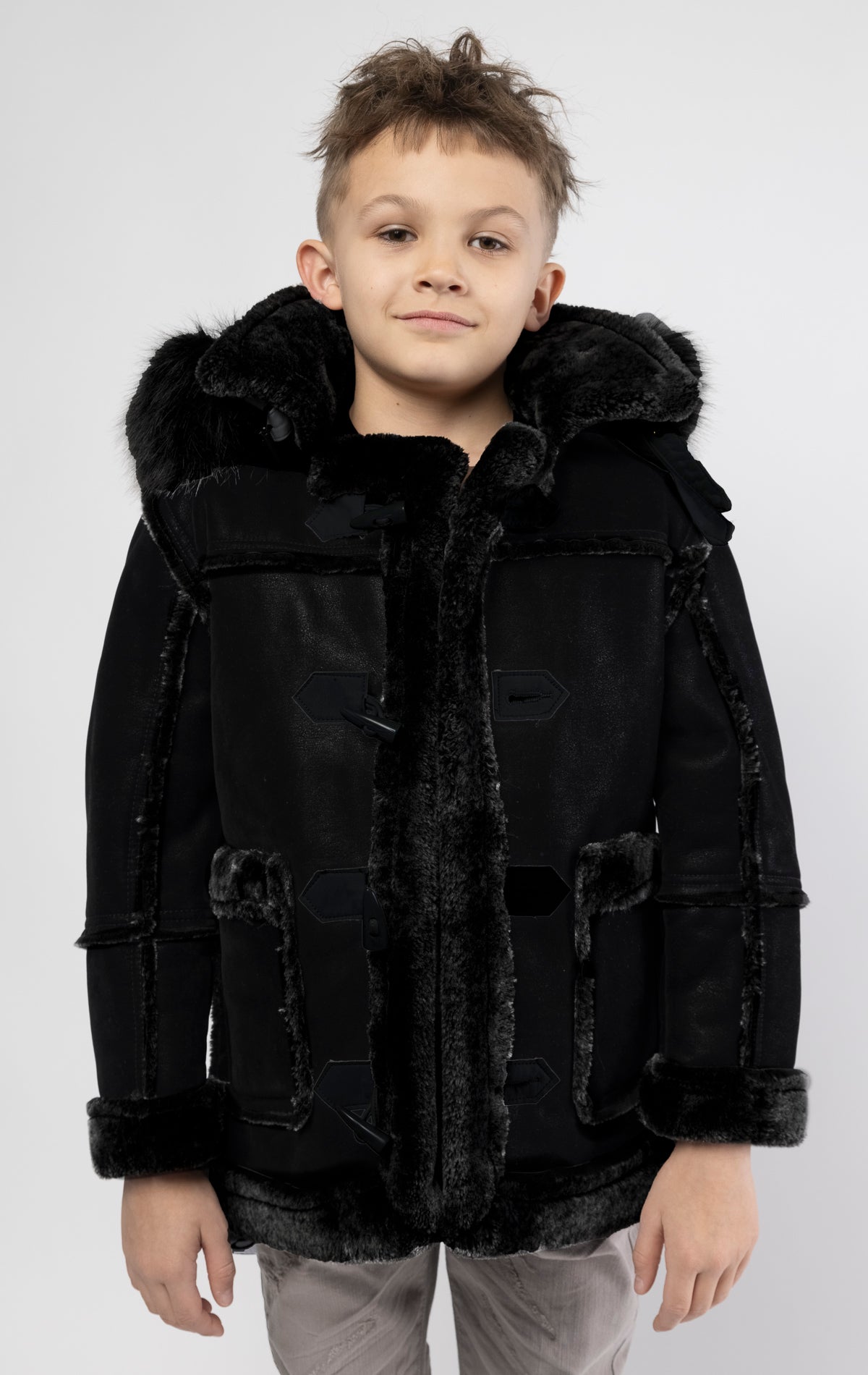 Black jacket lined with plush faux shearling throughout the body and sleeves. It also features shearling-accented pockets, a horn toggle front closure, and vegan suede straps with a buckle at the neck. For added convenience, a detachable faux fox fur hood