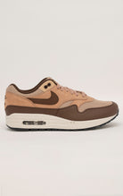 Nike Air Max 1 shoe in the "Cacao Wow" colorway. The shoe features deep brown tumbled leather on the mudguard and Swoosh, contrasted by tan nubuck overlays and a hairy suede tongue. The base mesh is a light brown color, and the midsole is crisp white. The outsole is black.