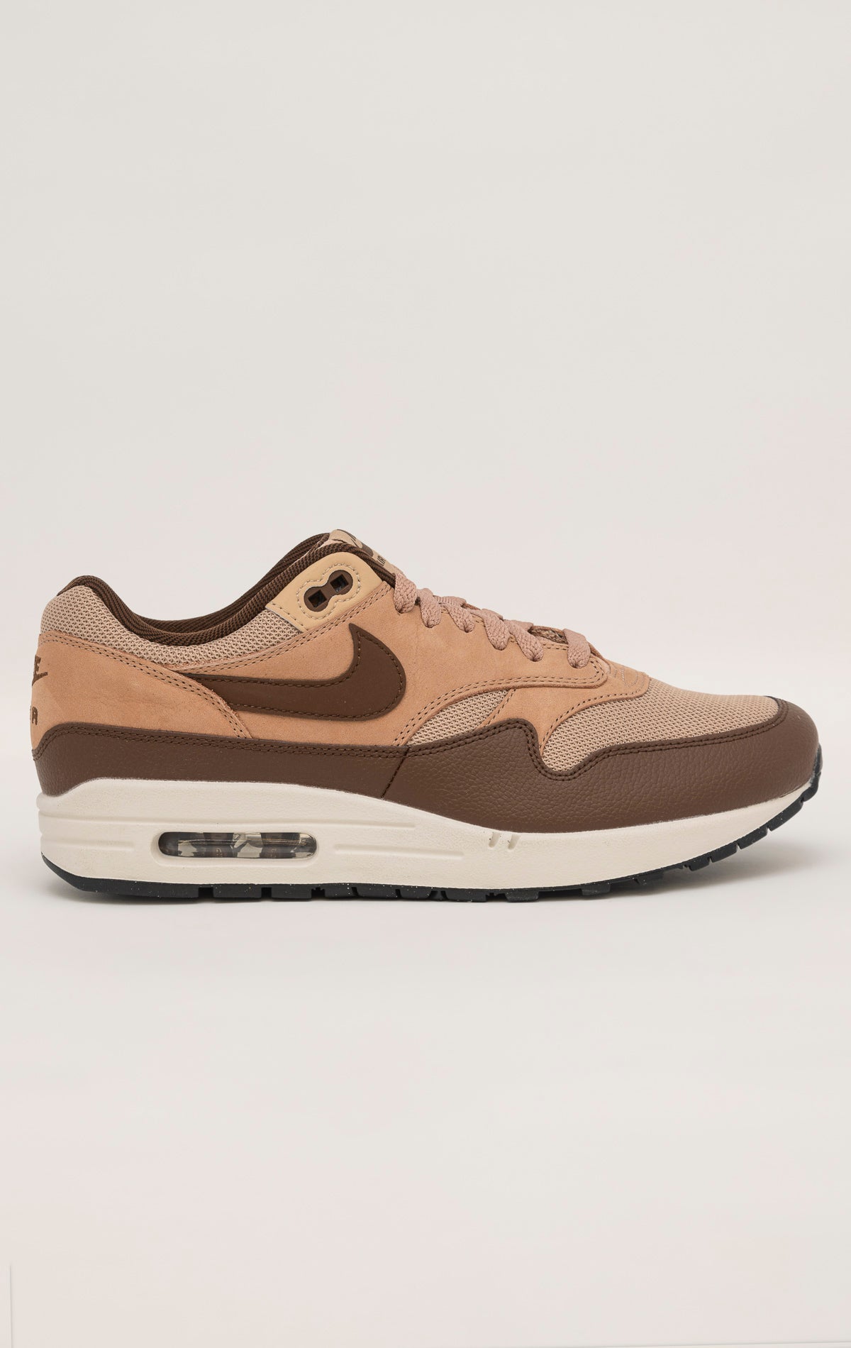 Nike Air Max 1 shoe in the "Cacao Wow" colorway. The shoe features deep brown tumbled leather on the mudguard and Swoosh, contrasted by tan nubuck overlays and a hairy suede tongue. The base mesh is a light brown color, and the midsole is crisp white. The