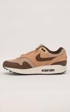 Nike Air Max 1 shoe in the "Cacao Wow" colorway. The shoe features deep brown tumbled leather on the mudguard and Swoosh, contrasted by tan nubuck overlays and a hairy suede tongue. The base mesh is a light brown color, and the midsole is crisp white. The outsole is black.