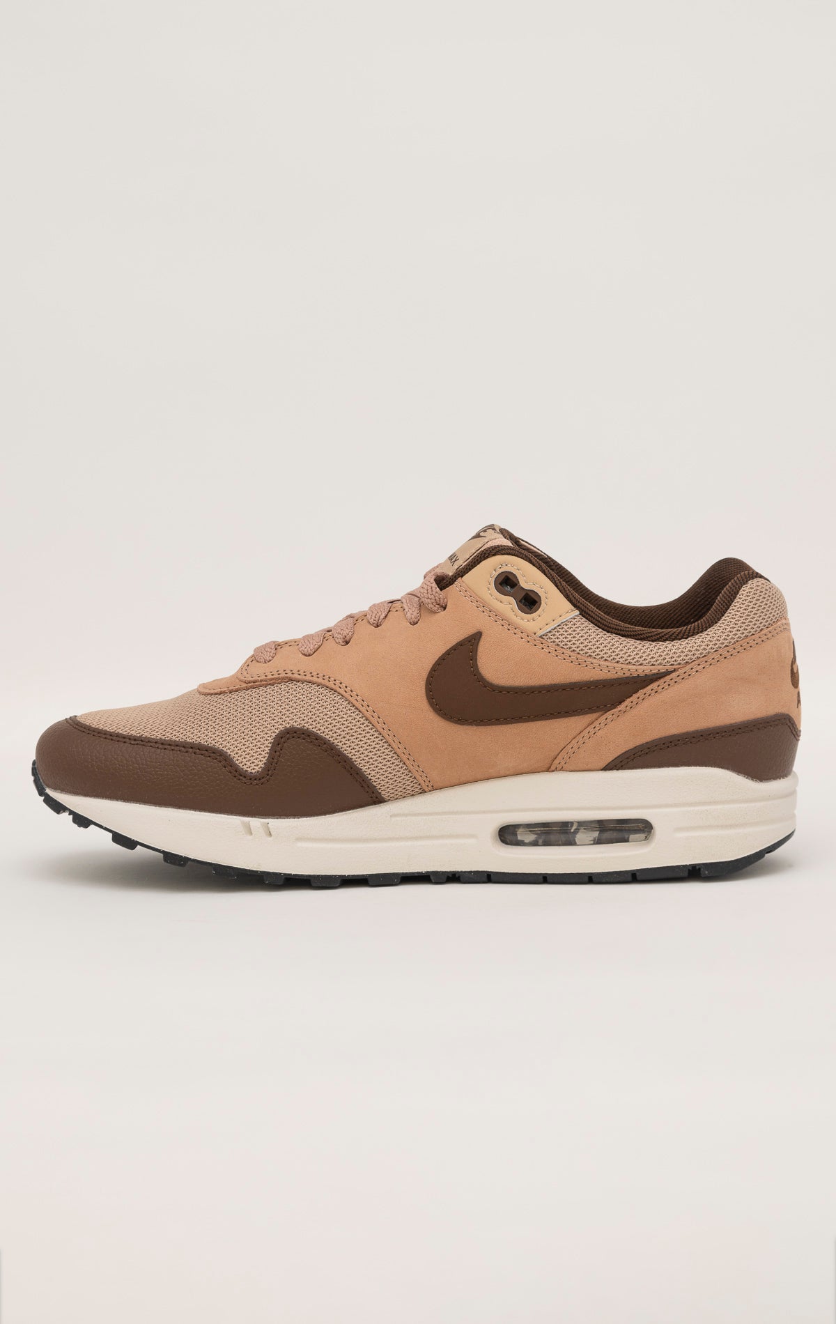 Nike Air Max 1 shoe in the "Cacao Wow" colorway. The shoe features deep brown tumbled leather on the mudguard and Swoosh, contrasted by tan nubuck overlays and a hairy suede tongue. The base mesh is a light brown color, and the midsole is crisp white. The