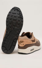 Nike Air Max 1 shoe in the "Cacao Wow" colorway. The shoe features deep brown tumbled leather on the mudguard and Swoosh, contrasted by tan nubuck overlays and a hairy suede tongue. The base mesh is a light brown color, and the midsole is crisp white. The outsole is black.