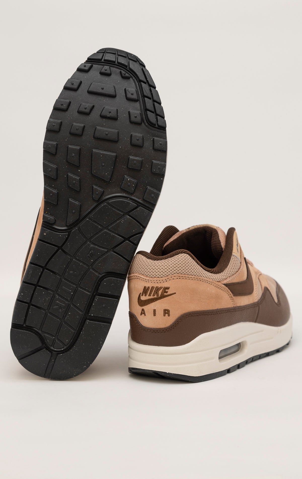 Nike Air Max 1 shoe in the "Cacao Wow" colorway. The shoe features deep brown tumbled leather on the mudguard and Swoosh, contrasted by tan nubuck overlays and a hairy suede tongue. The base mesh is a light brown color, and the midsole is crisp white. The