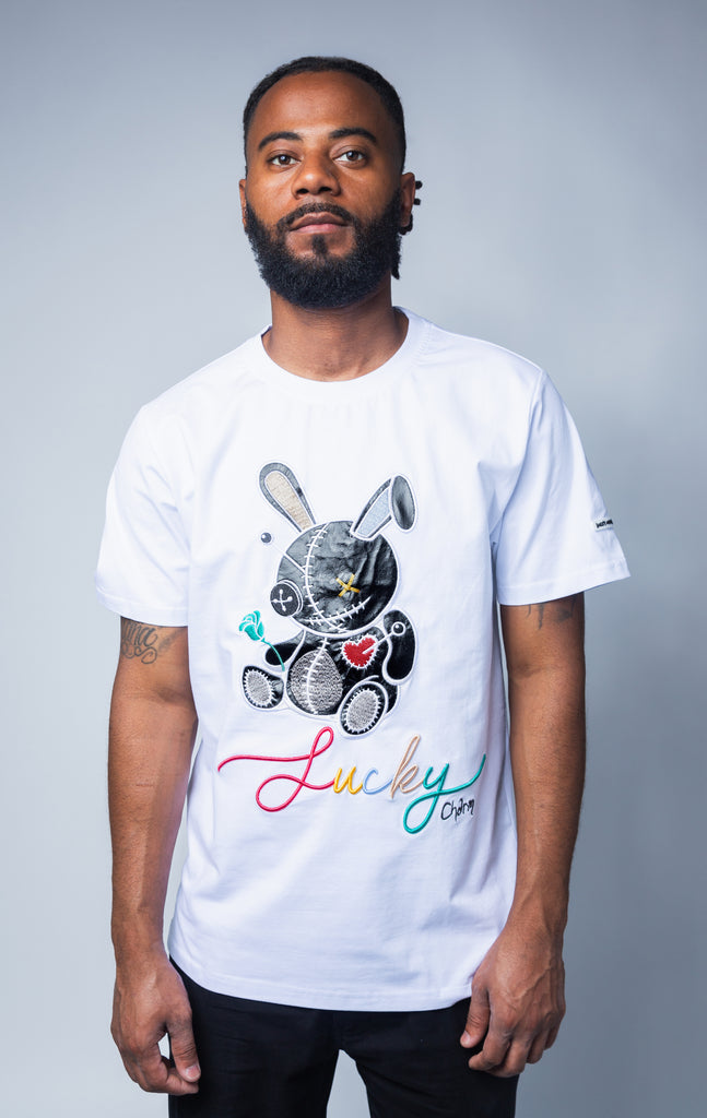 White crew neck  with bold Lucky Charm graphic print and applique on the front, short sleeves, rubber logo patch on the left sleeve, and square hem