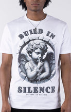 White t-shirt with a large graphic of a monochromatic cherub statue wearing chains and holding a finger to its lips. The graphic is surrounded by cracked stone and the text "BUILD IN SILENCE" is printed above and below in a textured black font.