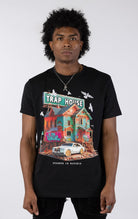 Black t-shirt with a large, colorful graphic of a run-down house with graffiti and a "TRAP HOUSE" sign on top. A white luxury car with gold rims is parked in front. White birds are flying around the house.