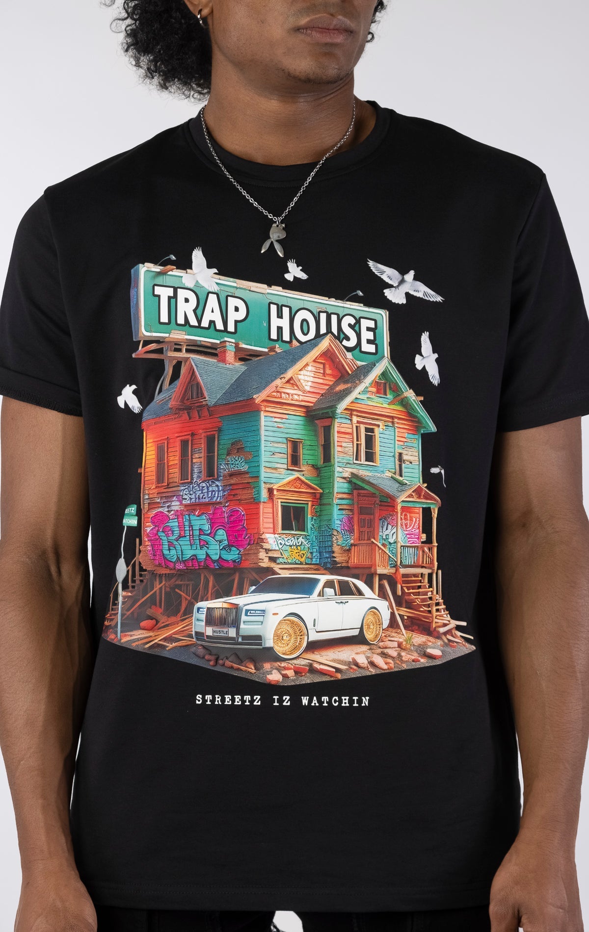 Black t-shirt with a large, colorful graphic of a run-down house with graffiti and a "TRAP HOUSE" sign on top. A white luxury car with gold rims is parked in front. White birds are flying around the house.