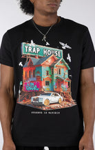 Black t-shirt with a large, colorful graphic of a run-down house with graffiti and a "TRAP HOUSE" sign on top. A white luxury car with gold rims is parked in front. White birds are flying around the house.