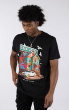 Black t-shirt with a large, colorful graphic of a run-down house with graffiti and a "TRAP HOUSE" sign on top. A white luxury car with gold rims is parked in front. White birds are flying around the house.