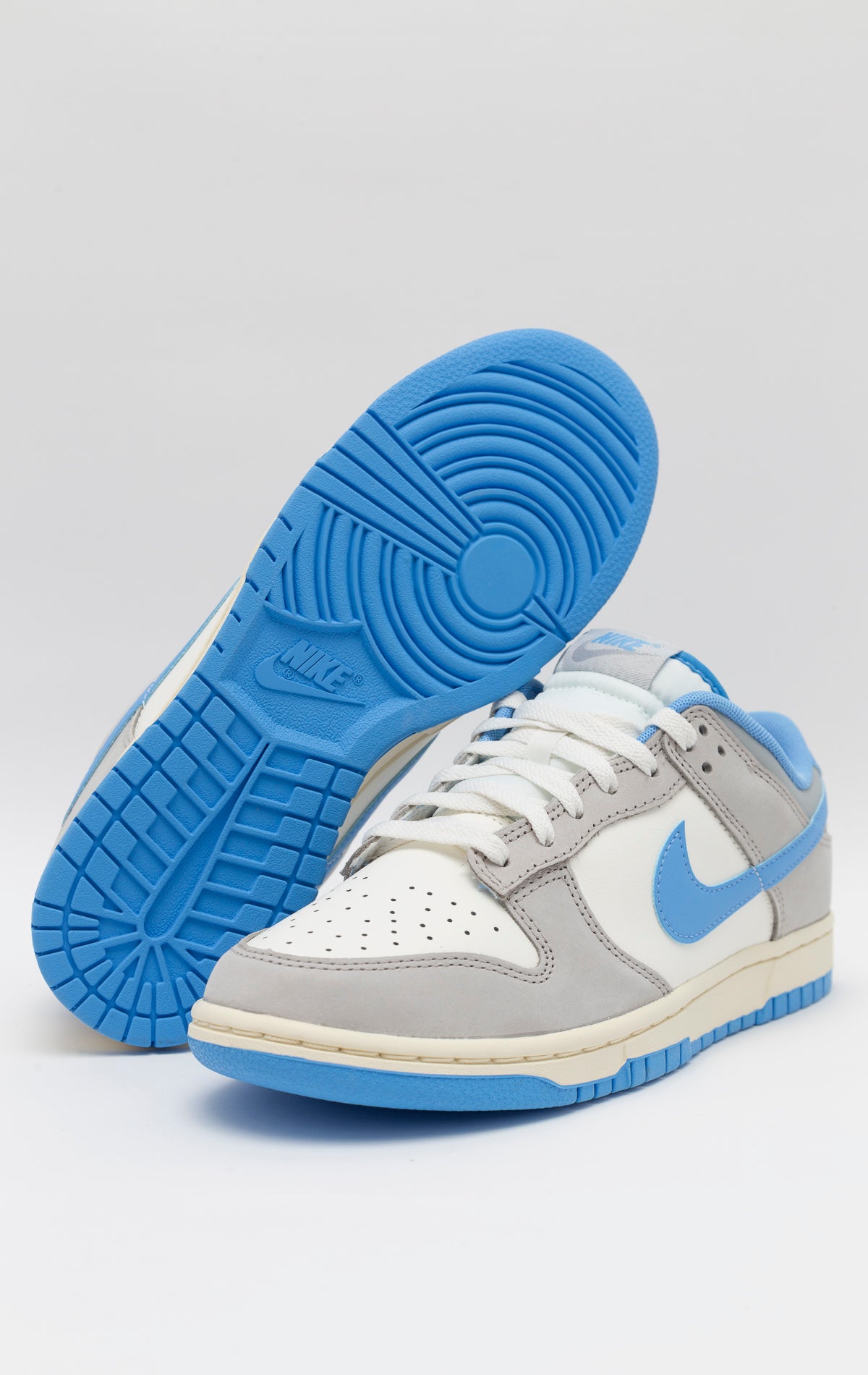 The Nike Dunk Low Athletic Department University Blue showcases a Light Smoke Gray upper with University Blue detailing. Made with high-quality leather and suede, it boasts both durability and fashion in a traditional low-top design. The padded collar, cushioned insole, and pivot point tread on the rubber outsole guarantee comfort. The Light Iron Ore EVA foam midsole offers impact defense. Iconic Swoosh logos can be found on both sides, and the tongue and heel tab feature Nike branding.