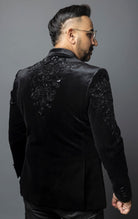 Black, slim-fit velvet blazer adorned with sequins and beads. The blazer has a peak lapel collar, long sleeves, two-button closure, and four-button cuffs. The blazer is made from a blend of 92% regenerated fabric and 8% spandex.