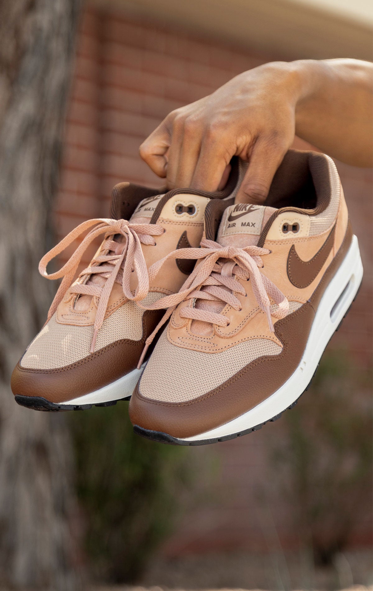 Nike Air Max 1 shoe in the "Cacao Wow" colorway. The shoe features deep brown tumbled leather on the mudguard and Swoosh, contrasted by tan nubuck overlays and a hairy suede tongue. The base mesh is a light brown color, and the midsole is crisp white. The outsole is black.