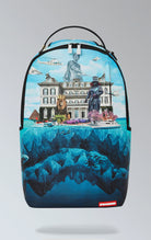 Eye-catching blue Sprayground backpack featuring artwork of a luxury mansion atop a shark, with separate laptop & sunglasses compartments, padded back, and luggage sleeve.