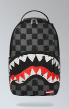 Gray Sprayground backpack with a checkered shark design. Features multiple compartments including a separate velour laptop compartment, side pockets, and a hidden zippered pocket. Equipped with ergonomic mesh back padding, adjustable straps, and a sliding back sleeve for attaching to carry-on luggage. Made from durable vegan leather.