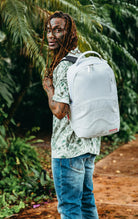 White Sprayground backpack with a unique scribble design. Features multiple compartments including a separate velour laptop compartment, side pockets, and a hidden zippered pocket. Equipped with ergonomic mesh back padding, adjustable straps, and a sliding back sleeve for attaching to carry-on luggage. Made from durable vegan leather.